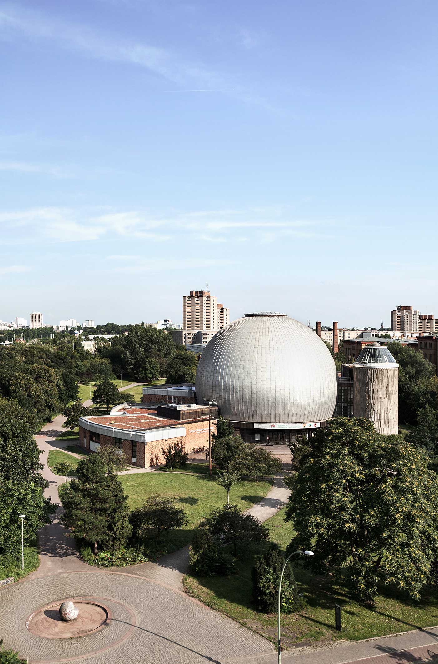 Das Zeiss-Großplanetarium in einem grünen Park, mit seiner markanten, kuppelförmigen Struktur. Im Hintergrund sind hohe Gebäude und ein klarer blauer Himmel sichtbar.