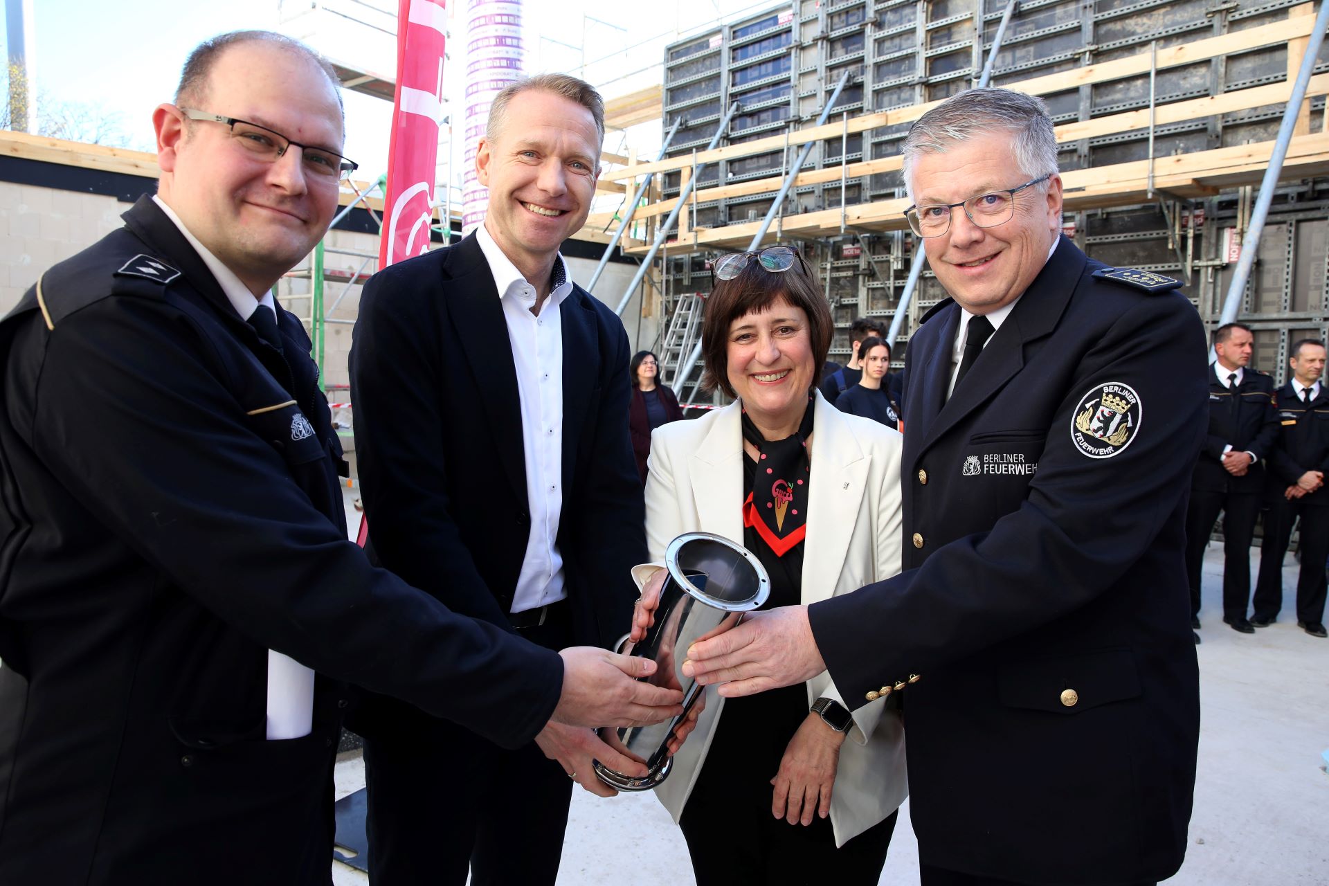 Wichtige Vertreter:innen der BIM und der Berliner Feuerwehr posieren mit einer Zeitkapsel vor einer Baustelle.