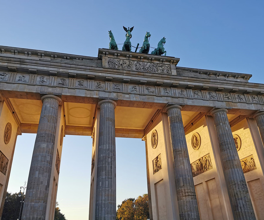 Das Brandenburger Tor in Berlin, von unten fotografiert, mit der Quadriga oben und den Säulen im Vordergrund, beleuchtet vom Sonnenlicht. Der Himmel ist klar und blau.