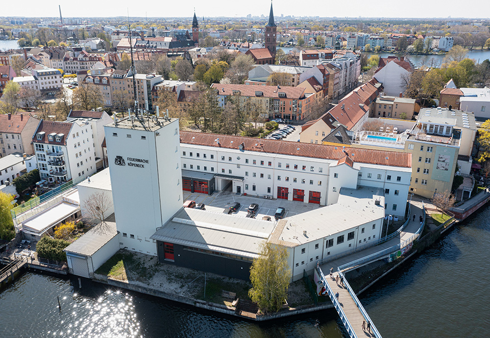 Eine Luftaufnahme der Feuerwache Köpenick, ein großes weißes Gebäude mit roten Toren, das von Wasser umgeben ist. Eine Brücke führt zum Gebäude, und im Hintergrund ist die Stadt mit verschiedenen Gebäuden und einer Kirche zu sehen.