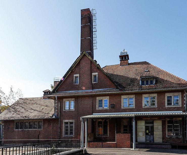 Historisches Backsteingebäude mit hohem Schornstein und roten Ziegeldächern auf der Liegenschaft „Am Stener Berg“. Die ehemalige Bäckerei zeigt markante Fassadendetails wie Rundbogenfenster und ein Vordach über dem Eingangsbereich, davor ein leerer Vorplatz bei klarem Himmel.