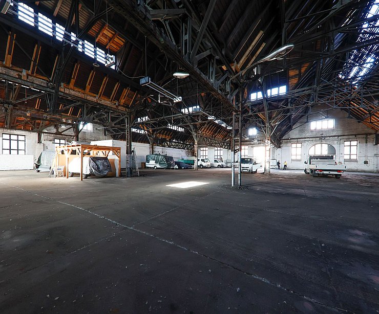 Große Industriehalle mit offenem Dachstuhl aus Holz, durch dessen Fensteröffnungen Licht einfällt. Der Boden ist asphaltiert, mehrere Fahrzeuge stehen am Rand, darunter ein Transporter und ein Pkw. Die Halle wirkt leer und bietet viel Platz.
