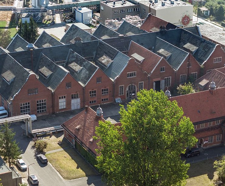 Luftaufnahme des denkmalgeschützten Gebäudeensembles „Am Stener Berg“ mit roten Backsteinfassaden und giebelständigen Dächern. Die Baukörper stehen dicht aneinandergereiht, davor ein kleiner Parkplatz mit Fahrzeugen und Bäume, im Hintergrund moderne Gewerbe- und Technikbauten.