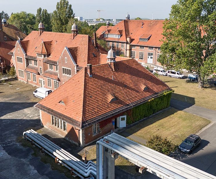 Ziegelrote Gebäude auf dem Gelände „Am Stener Berg“, aufgenommen aus erhöhter Perspektive. Die Dächer sind mit roten Ziegeln gedeckt, zwischen den Gebäuden befinden sich begrünte Flächen und gepflasterte Wege. Rechts parken mehrere Autos, im Hintergrund sind Bäume und weitere Gebäude sichtbar.