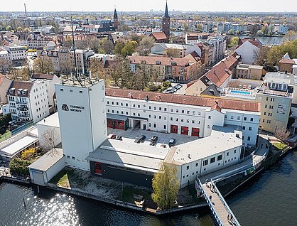 Eine Luftaufnahme der Feuerwache Köpenick, ein großes weißes Gebäude mit roten Toren, das von Wasser umgeben ist. Eine Brücke führt zum Gebäude, und im Hintergrund ist die Stadt mit verschiedenen Gebäuden und einer Kirche zu sehen.