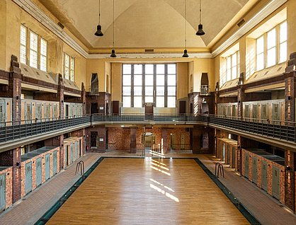 Innenansicht des Hubertus Bads mit hohen Decken und großen Fenstern. Der Raum ist symmetrisch gestaltet und hat mehrere Reihen von Umkleidekabinen mit Holzverkleidung. Die Wände und die Decke sind in warmen Gelbtönen gehalten.