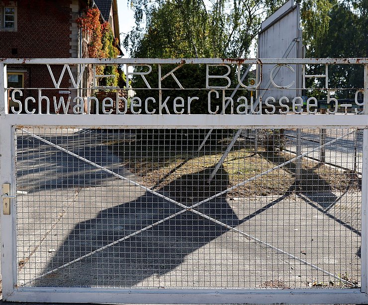 Weiß lackiertes Metalltor mit dem Schriftzug „Werk Buch – Schwanebecker Chaussee 5–9“. Das Gittertor ist geometrisch gestaltet und steht vor einem gepflasterten Hof. Im Hintergrund sind rote Backsteingebäude sowie Bäume zu sehen – Teil der Liegenschaft „Am Stener Berg“ in Berlin-Buch.