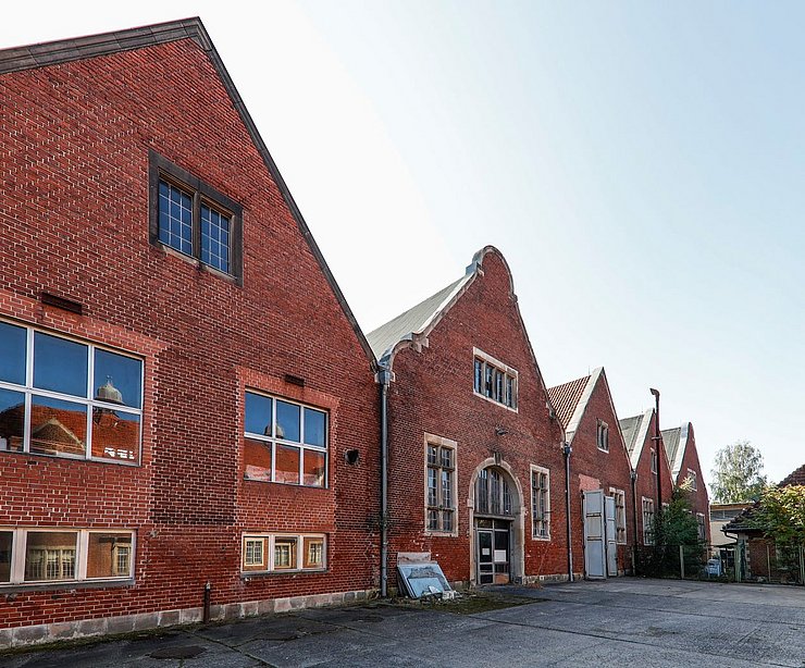 Backsteinbau mit giebelständiger Architektur auf der Liegenschaft „Am Stener Berg“, vermutlich eine ehemalige Fahrzeughalle. Mehrere große Fensterreihen an der Fassade, davor ein leerer Hof mit Kiesboden, aufgenommen bei klarem Himmel und Tageslicht.