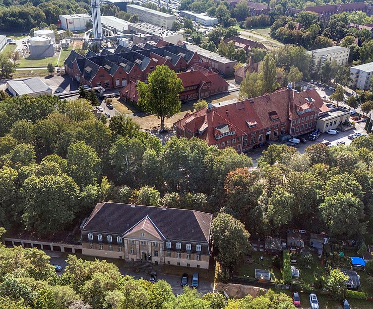 Luftaufnahme der Liegenschaft „Am Stener Berg“ in Berlin-Buch. Im Zentrum des Bildes befinden sich mehrere denkmalgeschützte Backsteingebäude mit roten Ziegeldächern, umgeben von dichtem Baumbestand. Im Vordergrund ist ein kleinerer Bau mit Walmdach zu sehen, im Hintergrund erstrecken sich weitere Gebäude und Straßen. Die Anlage wirkt eingebettet in eine grüne, ruhige Umgebung.