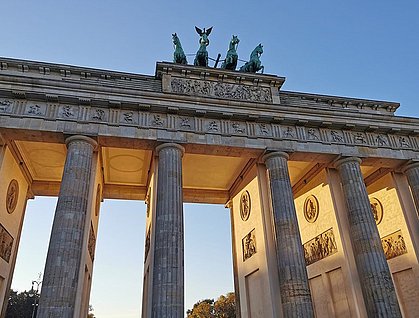 Das Brandenburger Tor in Berlin, von unten fotografiert, mit der Quadriga oben und den Säulen im Vordergrund, beleuchtet vom Sonnenlicht. Der Himmel ist klar und blau.